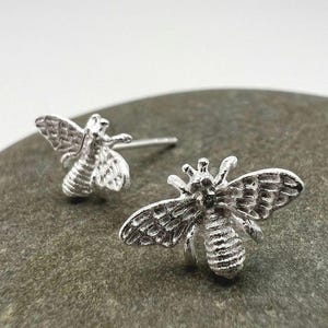 Peut inclure: Une paire de boucles d'oreilles en forme d'abeille argentées. Les boucles d'oreilles, avec des ailes et des segments de corps détaillés, reposent sur une surface en pierre grise. Bijoux en forme d'abeille, accessoire inspiré de la nature.