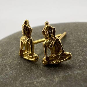 May include: A pair of gold-coloured Buddha stud earrings. Each earring depicts a small, detailed sculpture of a seated Buddha in a meditative pose. The earrings are set against a neutral background, highlighting their intricate design and metallic sheen.