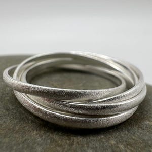 Könnte beinhalten: Ein Satz von fünf ineinandergreifenden silbernen Ringen mit einer strukturierten, matten Oberfläche. Die Ringe sind ineinander verschlungen und erzeugen einen geschichteten Effekt. Der Schmuck wird vor einem neutralen Hintergrund präsentiert, der den metallischen Glanz hervorhebt.