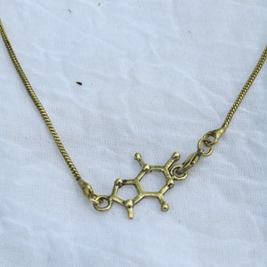 Op de afbeelding: Een gouden ketting met een hanger in de vorm van een chemische structuur. De hanger is gemaakt van metaal en heeft een glanzende afwerking.