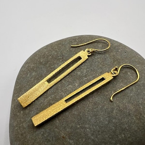 May include: A pair of gold-colored earrings with a rectangular design and a cutout in the center. The earrings have a textured surface and are displayed on a gray stone. The hooks are curved.
