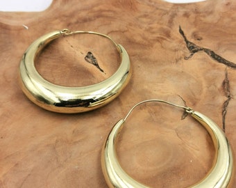 May include: Two gold-colored hoop earrings. The earrings have a thick, rounded design and a shiny finish. They are displayed on a wooden surface with a natural grain pattern.