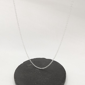 Puede incluir: Un delicado collar de cadena plateada descansa sobre una superficie redonda, oscura y texturizada. El collar es sencillo, con una cadena fina y un diseño sutil y elegante. El fondo es blanco y brillante, resaltando los detalles del collar.