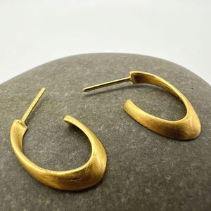 Könnte beinhalten: Ein Paar goldfarbene Creolen. Die Ohrringe haben eine glatte, matte Oberfläche und eine geschwungene, ovale Form. Die Stifte sind gerade und erstrecken sich von der Oberseite der Creolen. Die Ohrringe werden auf einer grauen Steinoberfläche präsentiert.