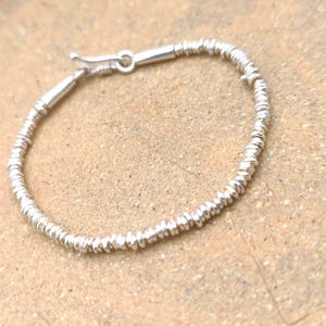 May include: A silver-toned bracelet composed of numerous small, thin, disc-shaped beads strung together. The bracelet features a smooth, cylindrical metal clasp mechanism at one end, allowing for a secure closure of the circular jewelry piece.