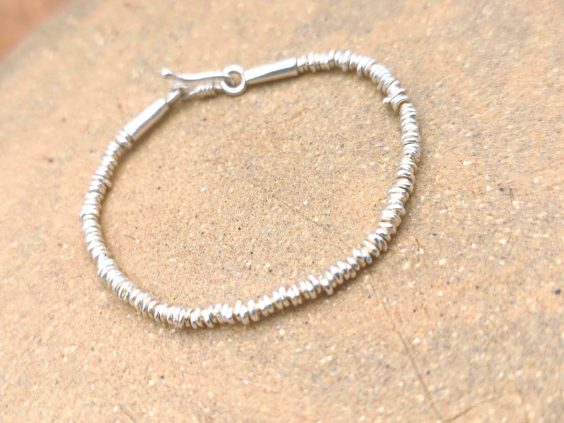 May include: A silver-toned bracelet composed of numerous small, thin, disc-shaped beads strung together. The bracelet features a smooth, cylindrical metal clasp mechanism at one end, allowing for a secure closure of the circular jewelry piece.