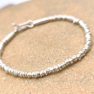 May include: A silver bracelet composed of numerous small, thin, disc-shaped beads strung together in a continuous circular pattern. The bracelet features a metallic finish and includes a small, functional silver hook-and-eye clasp mechanism.