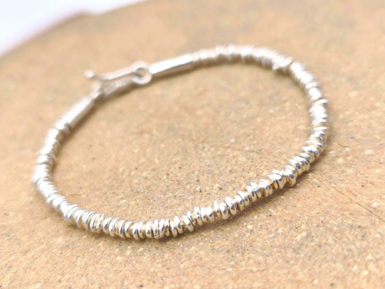 May include: A silver bracelet composed of numerous small, thin, disc-shaped beads strung together in a continuous circular pattern. The bracelet features a metallic finish and includes a small, functional silver hook-and-eye clasp mechanism.