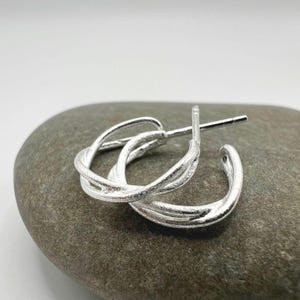 Könnte beinhalten: Ein Paar silberne Creolen mit einem gedrehten Design. Die Ohrringe sind aus glänzendem Metall gefertigt und haben einen Steckerverschluss. Die Reifen sind klein und kreisförmig, mit einer strukturierten Oberfläche.