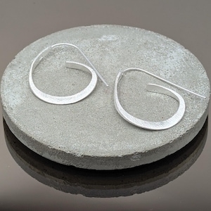 Puede incluir: Un par de pendientes plateados, de aro abierto, con un acabado de metal cepillado. Los pendientes tienen un diseño asimétrico único, apoyados sobre una superficie gris y circular. Los ganchos se extienden hacia afuera.