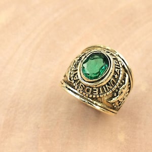 May include: A gold-colored ring with a large, oval-shaped green gemstone at its center. The ring is engraved with the words "UNITED STATES AIR FORCE" and other details. The ring is set against a light brown background.