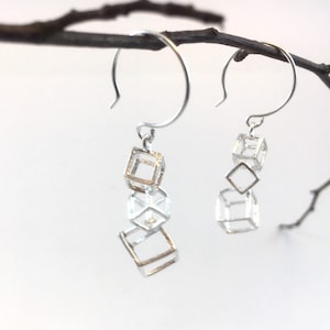 Könnte beinhalten: Silberne Hängeohrringe mit geometrischen Würfel- und Rauteformen. Die Ohrringe hängen an silbernen Haken und weisen eine Mischung aus silber- und goldfarbenem Metall auf. Das Design ist modern und minimalistisch, mit klaren Linien und offenen Räumen.