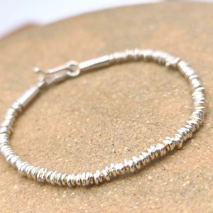 Könnte beinhalten: Ein silbernes Armband, das aus zahlreichen kleinen, dünnen, scheibenförmigen Perlen besteht, die aneinandergereiht sind. Das Armband hat eine metallisch silberne Oberfläche und einen kleinen, funktionalen Hakenverschluss zum Schließen.