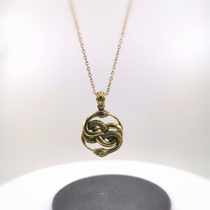 Puede incluir: Un collar con colgante dorado con un diseño circular de serpientes entrelazadas. Las serpientes están detalladas con escamas y cabezas mirando hacia afuera. La cadena del collar es delicada y dorada, con un cierre sencillo.