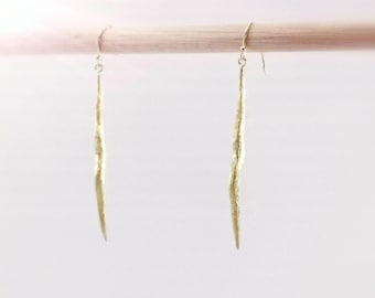 Puede incluir: Dos pendientes alargados de color dorado con superficie texturizada cuelgan de una clavija de madera sobre un fondo blanco. Los pendientes tienen una forma delgada y orgánica, que se estrecha hasta un punto. Los ganchos son plateados.