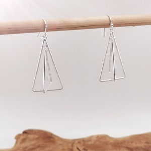 Könnte beinhalten: Ein Paar silberne Dreiecksohrringe mit geometrischem Design. Die Ohrringe hängen an einem Holzstab.