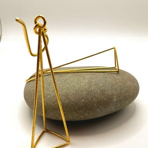 Könnte beinhalten: Goldfarbene geometrische Ohrringe. Ein Ohrring hat eine dreieckige Form mit einem Haken, der andere ein lineares Design. Die Ohrringe werden auf einem grauen Stein vor weißem Hintergrund präsentiert. Die Ohrringe sind aus Metall.