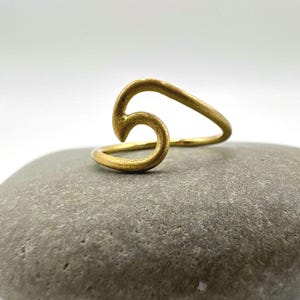 Op de afbeelding: Een goudkleurige ring met een golfachtig ontwerp. De ring is gemaakt van glanzend metaal en ligt op een grijze steen. Het ontwerp van de ring heeft een gebogen, open vorm.