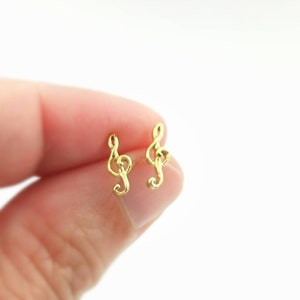 Puede incluir: Un par de pendientes pequeños dorados con forma de clave de sol. Los pendientes son de estilo botón, con un diseño sencillo y elegante. El fondo es blanco neutro.