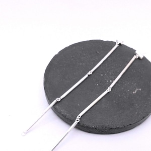 Puede incluir: Un par de pendientes lineales plateados con un diseño minimalista. Los pendientes se muestran sobre una piedra circular gris oscuro sobre un fondo blanco. Los pendientes tienen un estilo sencillo y elegante.