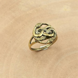 Può includere: Un anello color oro con un designo di due serpenti intrecciati. I serpenti sono riccamente dettagliati e creano un motivo a nodo.