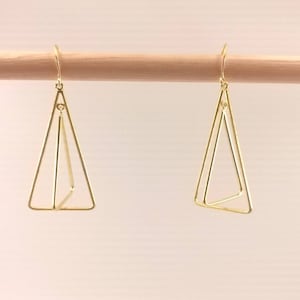 Può includere: Un paio di orecchini geometrici color oro. Ogni orecchino presenta un design triangolare a doppio strato sospeso a un gancio. Gli orecchini sono esposti su uno sfondo chiaro, evidenziando il loro stile delicato e minimalista.