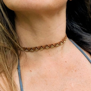 May include: A brown choker necklace with gold-coloured beads. The necklace is made of woven material and sits close to the neck. The beads are evenly spaced along the front of the necklace, adding a touch of elegance to the design.