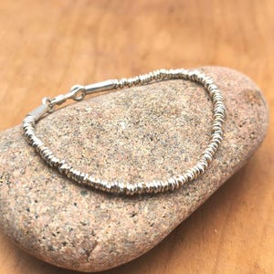 May include: A silver bracelet composed of numerous small, thin, disc-shaped beads strung together. The bracelet features a simple hook-and-eye clasp mechanism made of polished silver, creating a continuous circular design with a metallic finish.