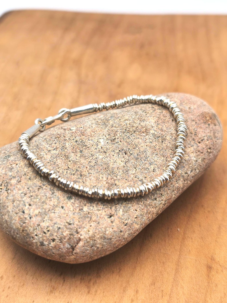 May include: A silver bracelet composed of numerous small, thin, disc-shaped beads strung together. The bracelet features a simple hook-and-eye clasp mechanism made of polished silver, creating a continuous circular design with a metallic finish.
