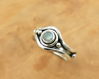 Op de afbeelding: Een zilveren ring met een maansteen in een bezel-setting. De ring heeft een uniek, golvend ontwerp en is versierd met kleine zilveren kralen.