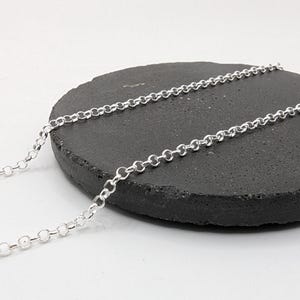 May include: A silver chain necklace with a round link design is displayed on a dark grey, textured, round stone. The necklace is laid across the stone and extends off the side. The background is white.
