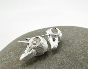 May include: A pair of silver-colored stud earrings, each shaped like a bird skull. The earrings have a detailed, textured surface and are set against a neutral background. The design is gothic and unique.