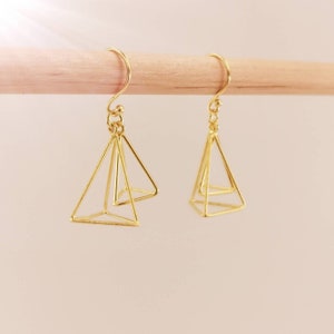 May include: A pair of gold-colored geometric earrings. Each earring features a wireframe pyramid design suspended from a curved ear wire. The earrings are displayed against a light background.