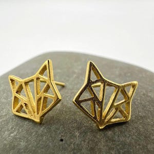 May include: A pair of gold-colored fox-shaped stud earrings. The earrings feature a geometric, openwork design, with the fox's head and ears clearly defined. The earrings are displayed on a neutral background.