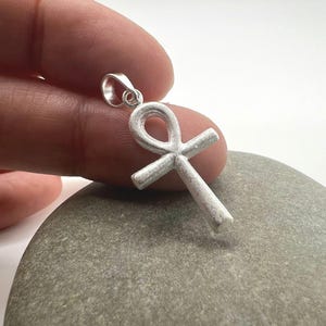 Könnte beinhalten: Ein silberner Ankh-Anhänger, ein Symbol des Lebens, wird gezeigt. Der Anhänger hat oben eine Schlaufe und unten eine Kreuzform. Der Anhänger wird vor einem hellen Hintergrund gehalten.