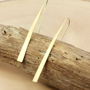 May include: A pair of gold-colored, linear drop earrings. Each earring features a long, flat, rectangular bar suspended from a curved ear wire. The earrings are displayed on a piece of driftwood.