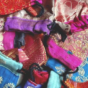 Puede incluir: Una colección de bolsas de tela coloridas en tonos de rojo, rosa, morado y azul. Las bolsas tienen cordones negros y están dispuestas sobre un fondo de tela estampada. Algunas bolsas tienen detalles dorados.