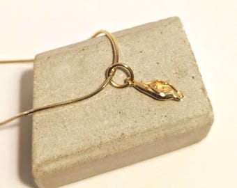 Puede incluir: Un collar de tono dorado con una cadena fina tipo serpiente y un pequeño colgante de forma orgánica e irregular. El colgante presenta una superficie texturizada y desigual con un acabado metálico, suspendido de una argolla circular.