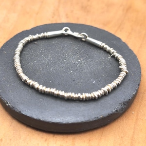 May include: A silver bracelet composed of numerous small, thin, disc-shaped beads strung together. The bracelet features a polished silver clasp mechanism at the top, connecting the two ends of the beaded strand into a circular shape.