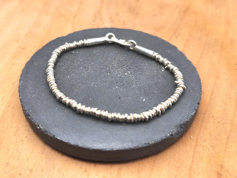 May include: A silver bracelet composed of numerous small, thin, disc-shaped beads strung together. The bracelet features a polished silver clasp mechanism at the top, connecting the two ends of the beaded strand into a circular shape.