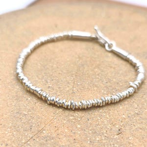 May include: A silver bracelet composed of numerous small, thin, disc-shaped metallic beads strung together in a continuous circular pattern, featuring a simple toggle clasp mechanism at the end for securing the piece around the wrist.