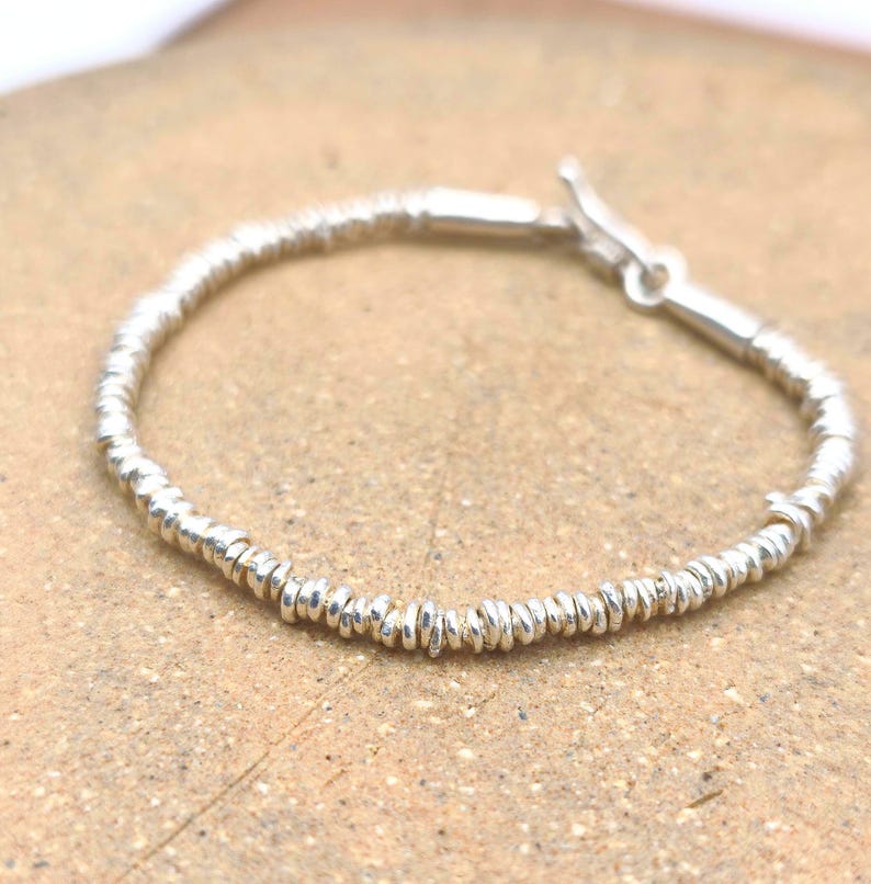 May include: A silver bracelet composed of numerous small, thin, disc-shaped metallic beads strung together in a continuous circular pattern, featuring a simple toggle clasp mechanism at the end for securing the piece around the wrist.