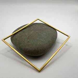 May include: A gold-coloured square bangle bracelet resting on a grey stone. The bracelet has a simple geometric design, with clean lines and a matte finish. The stone has a rough texture and a natural appearance.