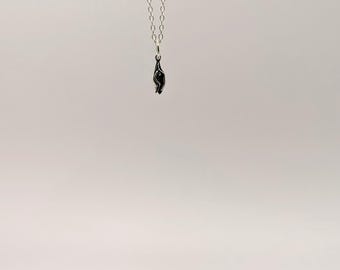 Puede incluir: Un collar de cadena de plata con un pequeño colgante negro en forma de mano. El collar cuelga de una base circular de hormigón gris.