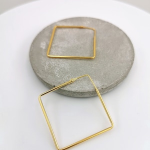 Könnte beinhalten: Zwei goldfarbene quadratische Creolen. Die Ohrringe werden auf einer grauen, kreisförmigen Oberfläche vor weißem Hintergrund präsentiert. Die Reifen sind geometrisch und modern gestaltet, mit einer schlichten, eleganten Ästhetik.