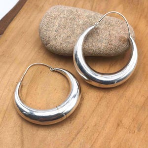 Può includere: Un paio di orecchini a cerchio in argento lucido con un design spesso, arrotondato e a forma di mezzaluna. Ogni orecchino presenta una sottile chiusura a filo che si aggancia alla parte superiore del cerchio, creando una superficie liscia.