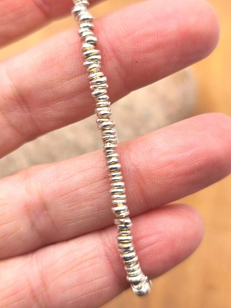May include: A single strand of small, metallic silver-colored beads arranged in a vertical line. Each bead features a slightly irregular, flattened circular shape with a reflective surface, creating a uniform texture along the entire length of the strand.