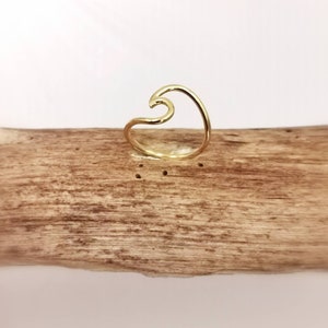 May include: A gold-colored ring in the shape of a wave. The ring is set on a piece of driftwood. The wave design is a continuous line, creating a minimalist and elegant look. The ring is a simple yet stylish piece of jewelry.