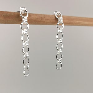 May include: A pair of silver chain earrings hanging from a wooden dowel rod. The earrings are made of a series of small, linked circles.