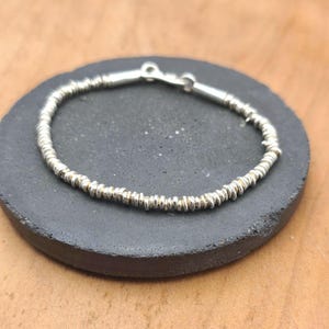 May include: A silver-toned bracelet composed of numerous small, thin, disc-shaped beads strung together in a continuous circular pattern. The bracelet features a simple metallic clasp mechanism at the top, connecting the two ends of the beaded strand.
